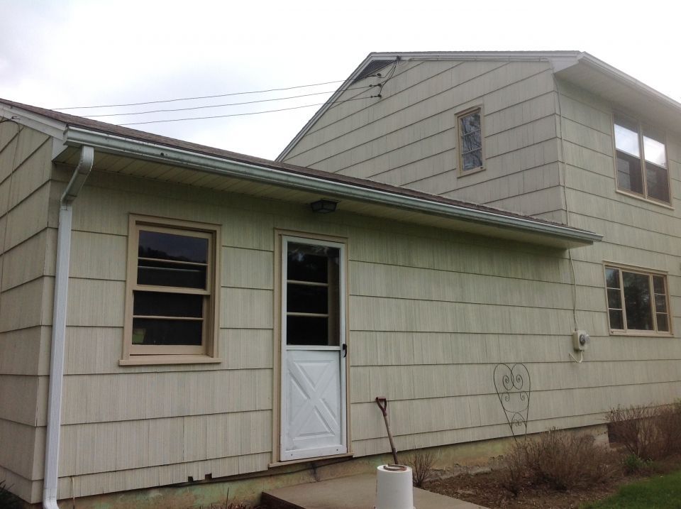 Side view of a two-story house with light siding. An attached single-story section has a door, window, and gutter.