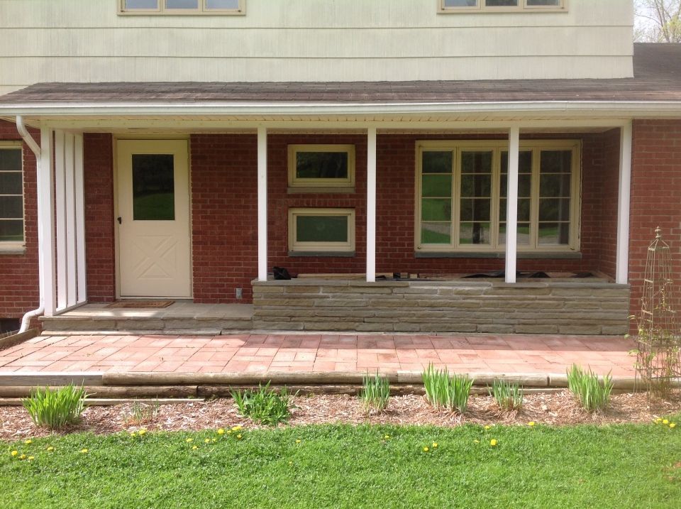 A brick house with a covered porch featuring a beige door, windows, and a brick and stone patio. A small garden borders the lawn.