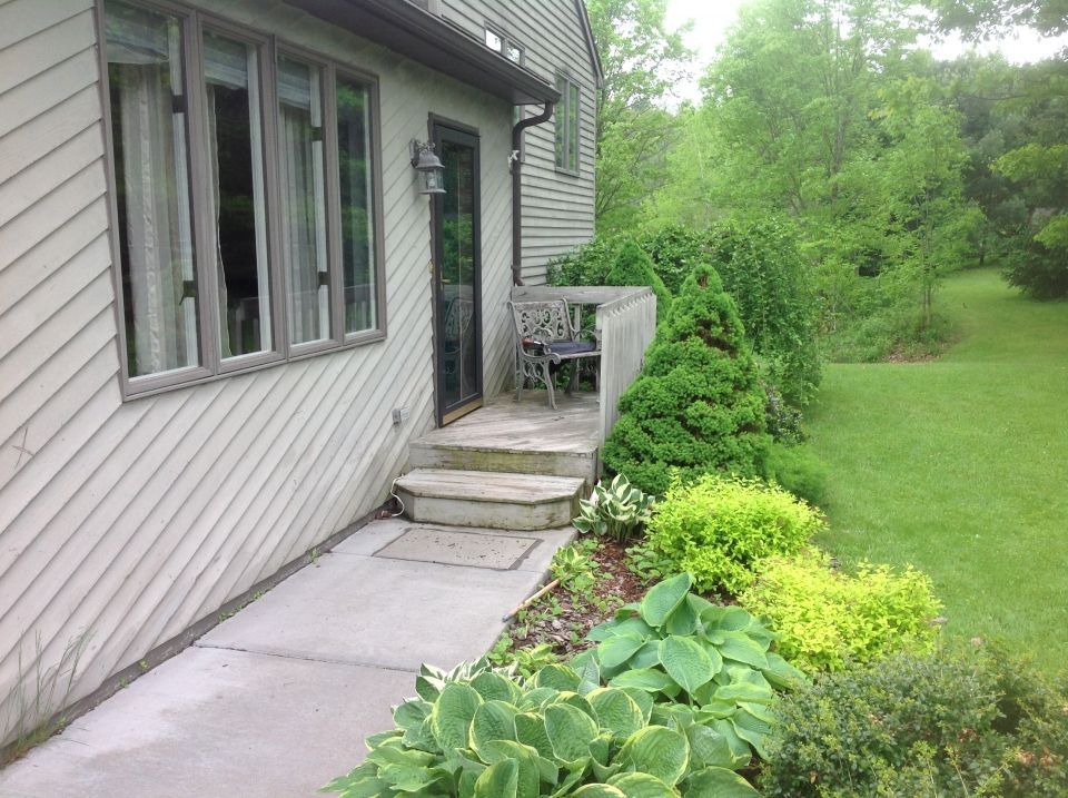 Exterior view of a house with a concrete walkway, steps, and a small patio area with a chair. Lush greenery borders the steps and lawn.