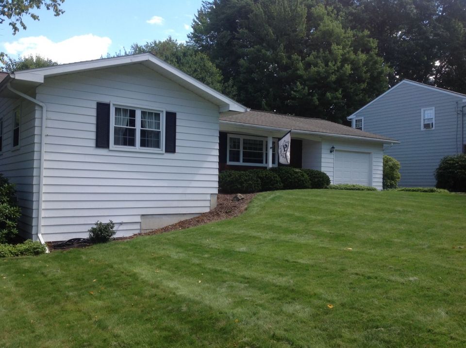 White house with black shutters, garage, and well-manicured lawn under a blue sky. A neighboring house is visible.