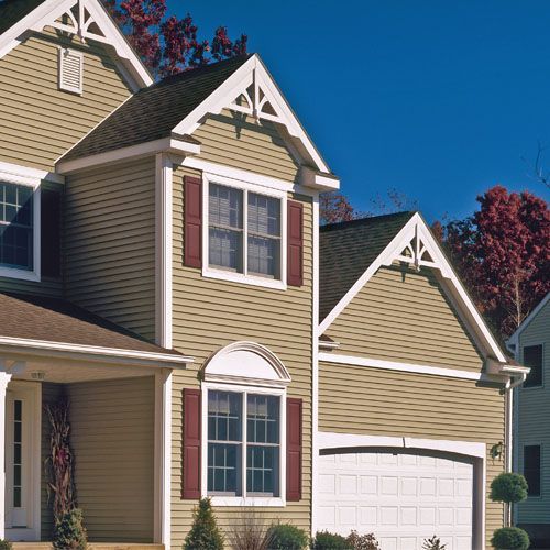 Two-story tan house with white trim, red shutters, and a white garage door, set against a blue sky.