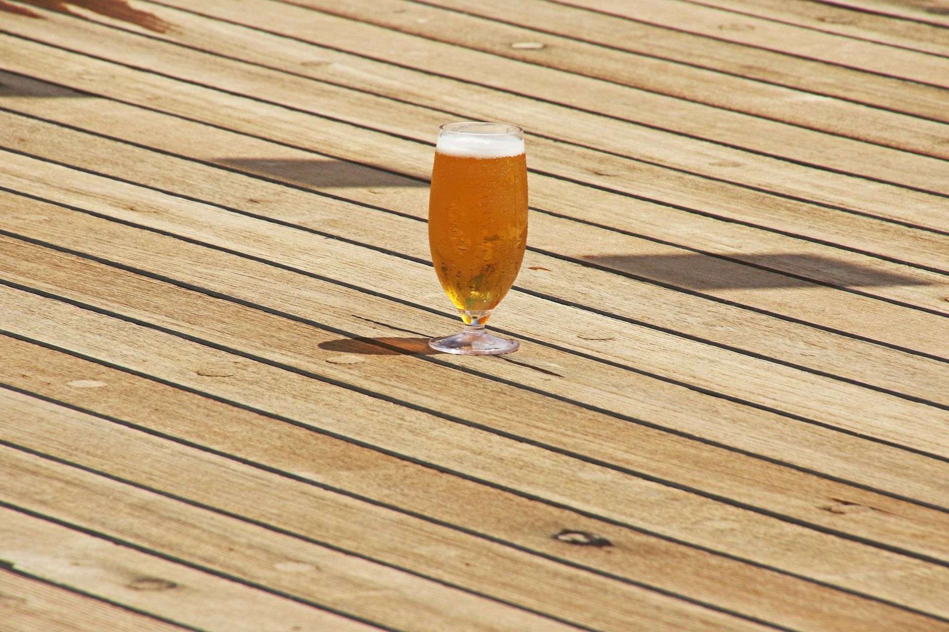 A glass of beer on a wooden deck, casting a shadow in sunlight.