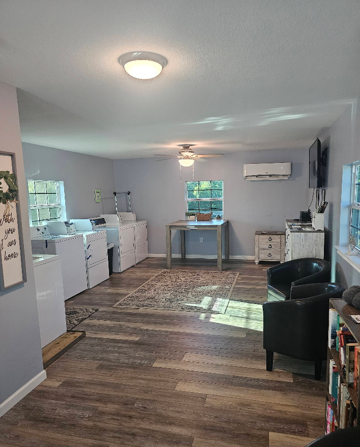 A laundromat interior with washing machines, tables, chairs, and a decorative rug on a wood floor.