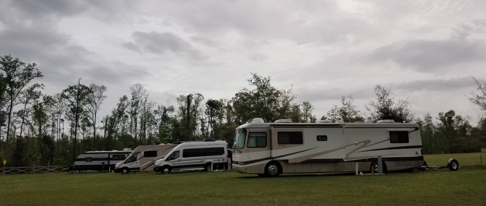Several RVs parked in a grassy field under a cloudy sky. Trees in the background.