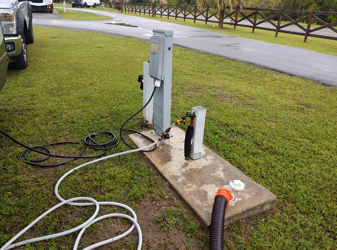 RV campsite hookups: electrical box, sewer connection, water hose, on a concrete pad.