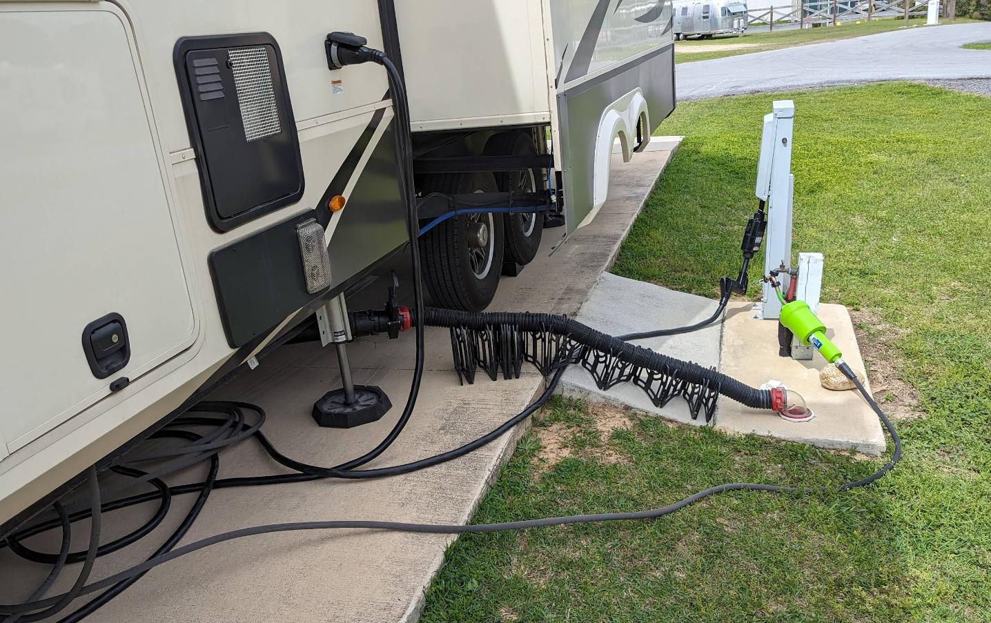 An RV connected to power and sewer at a campsite with cords on a concrete pad and grass.