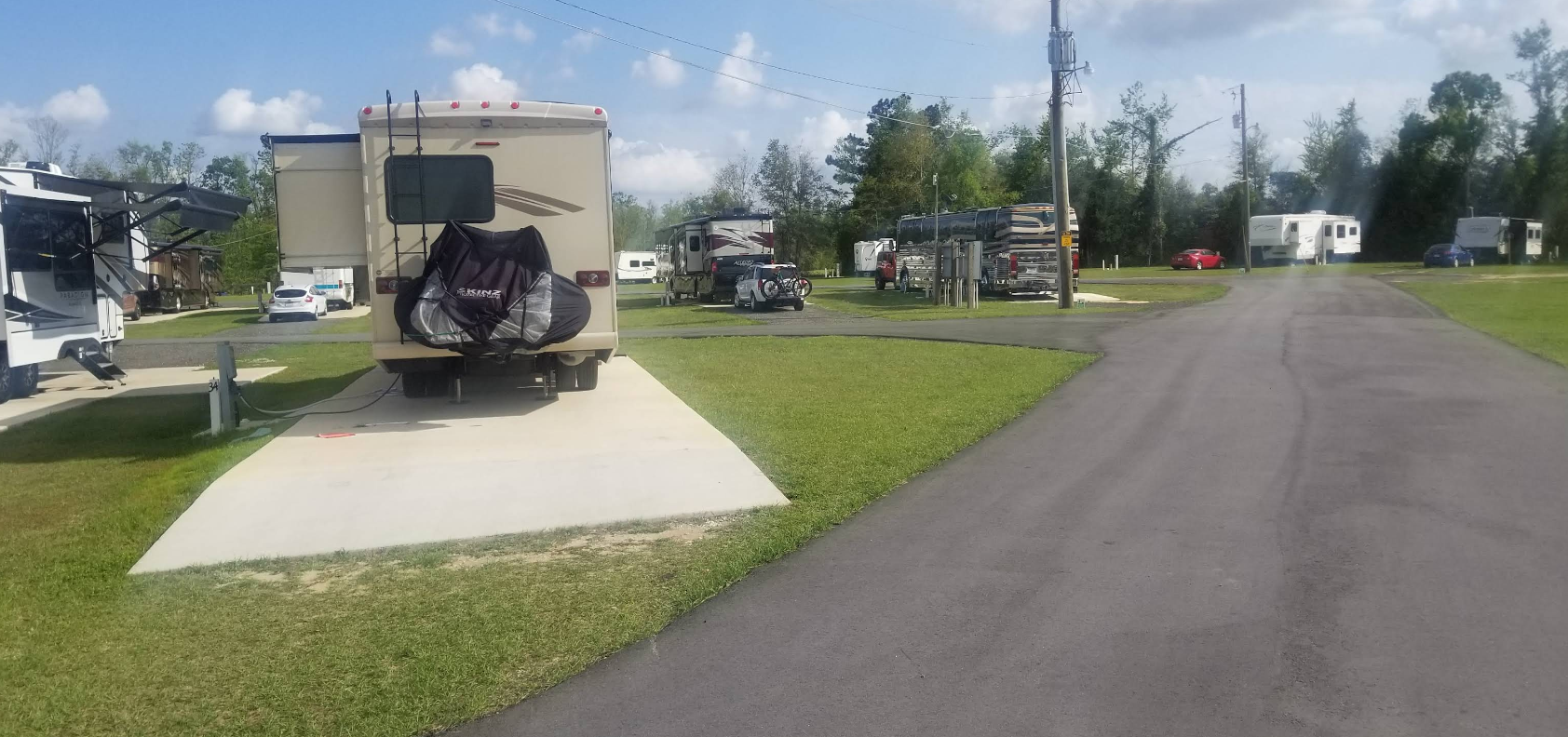 RV park with asphalt road, RVs parked on concrete pads, green grass, and a bright, blue sky.