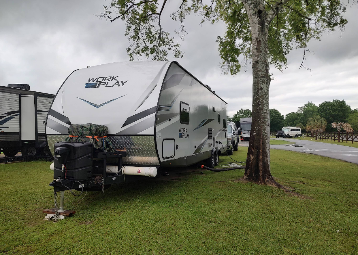 A travel trailer parked on a grassy campsite next to a tree. Overcast sky.