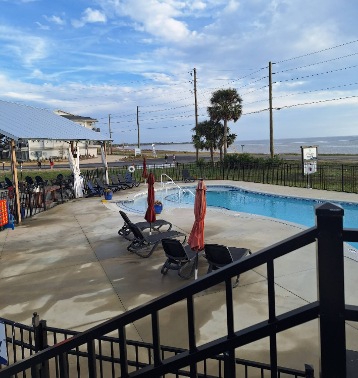 Poolside scene with lounge chairs, umbrellas, and a beach in the distance under a cloudy sky.