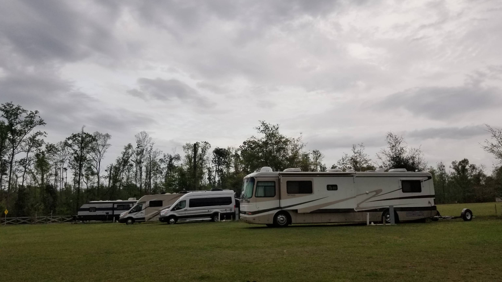 RVs and vans parked in a grassy field under a cloudy sky. Trees line the background.