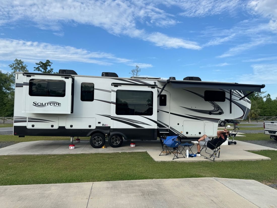 White and black RV parked on concrete pad with awning extended, person relaxing in chair.