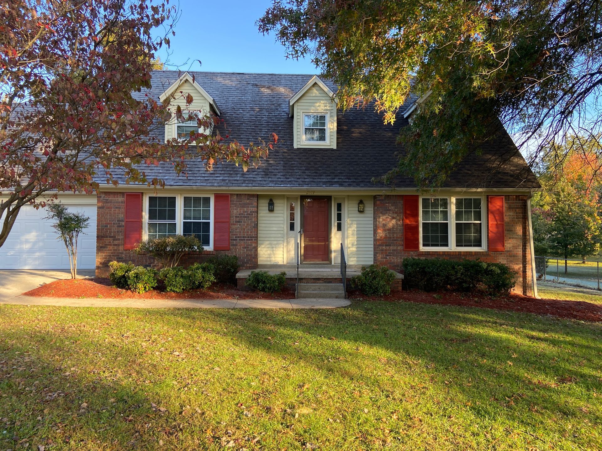 Brick house with red shutters, dormers, and a central door. Green lawn and fall trees in the yard.