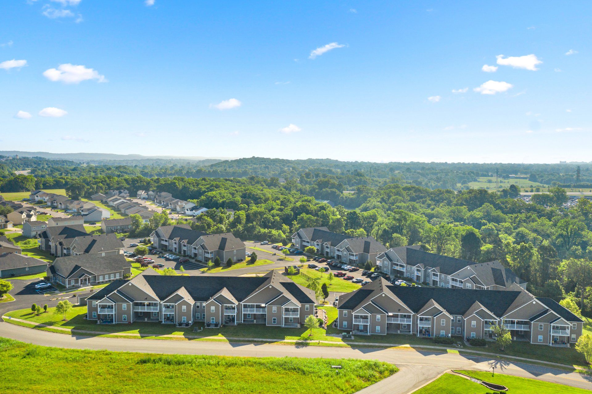 Aerial view of a residential area with gray apartment buildings surrounded by green fields and trees under a blue sky.