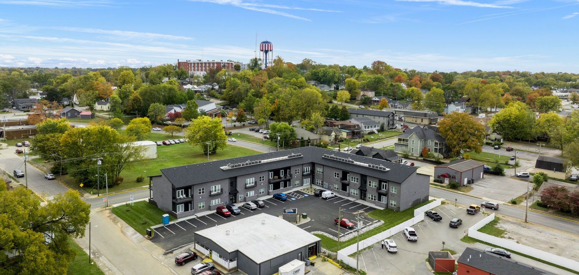 Aerial view of a gray apartment complex with a large parking lot, trees, and a water tower in the background.