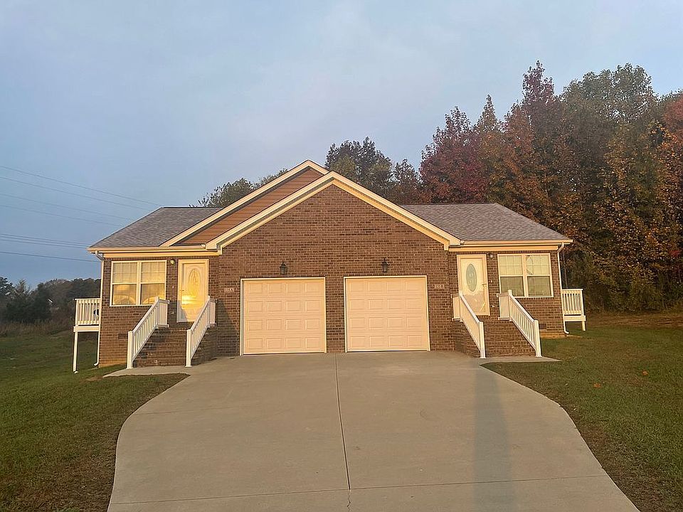 Brick duplex with two garage doors and front entrances. Concrete driveway and green lawn.