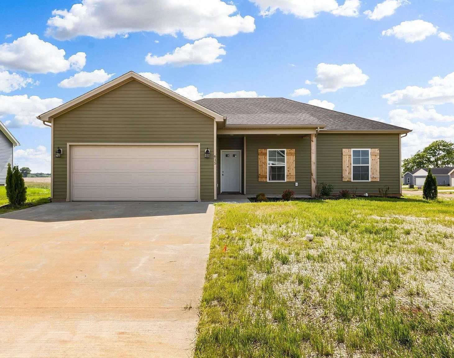 Green house with a two-car garage, concrete driveway, and grassy front yard under a blue sky.