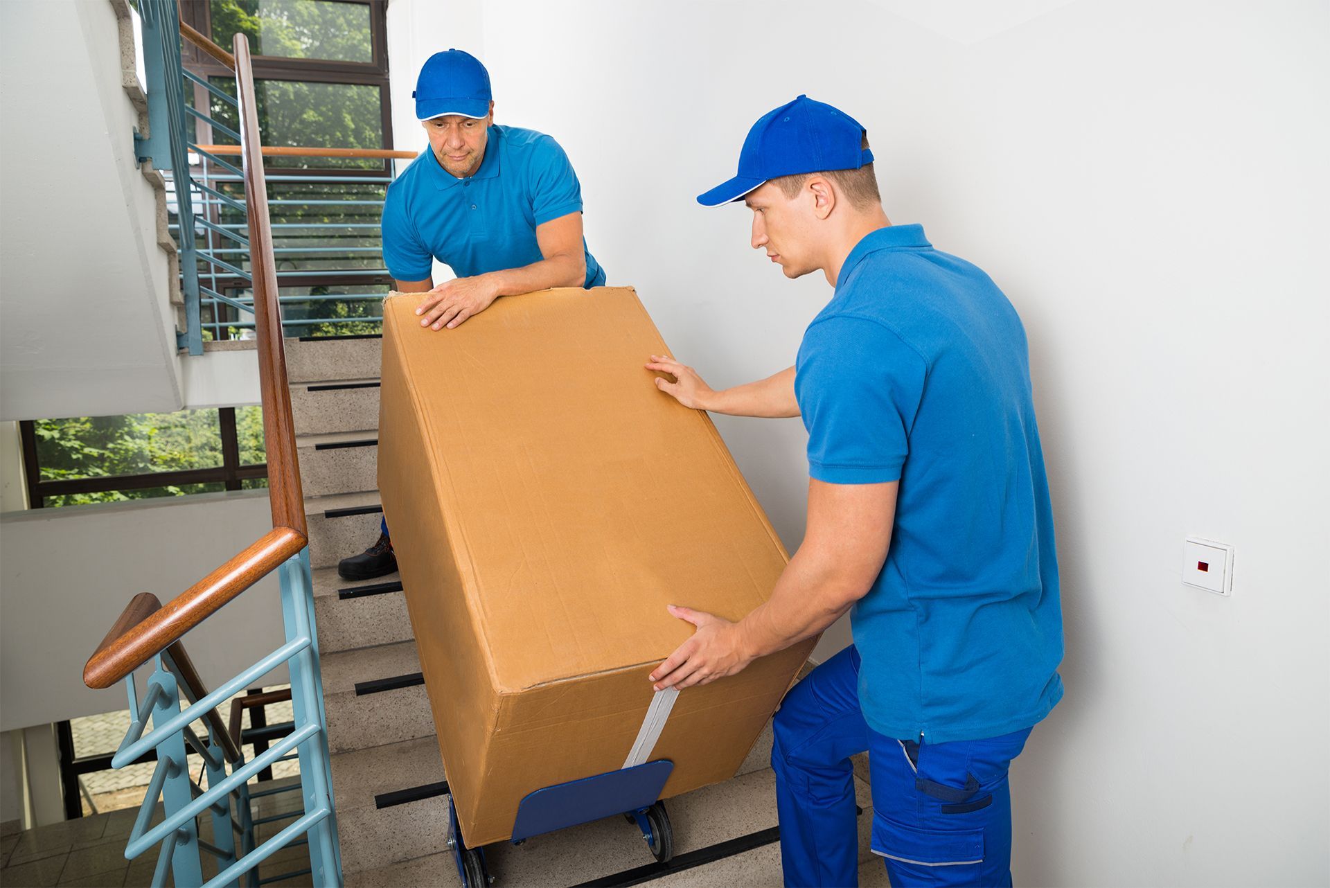 Movers Carrying a Cardboard Box Up a Set of Stairs