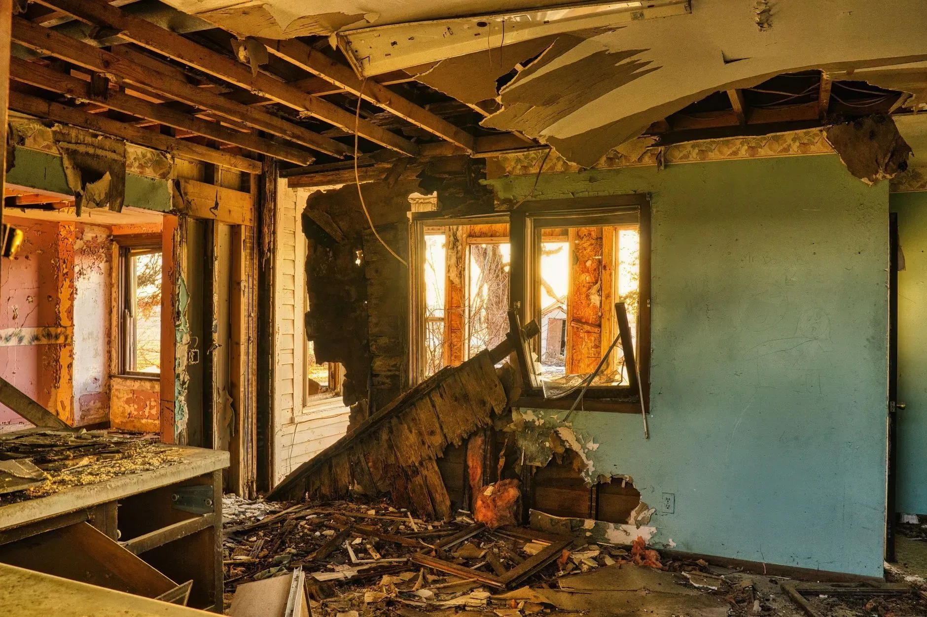Damaged room with exposed ceiling beams, broken walls, debris, and sunlight through windows