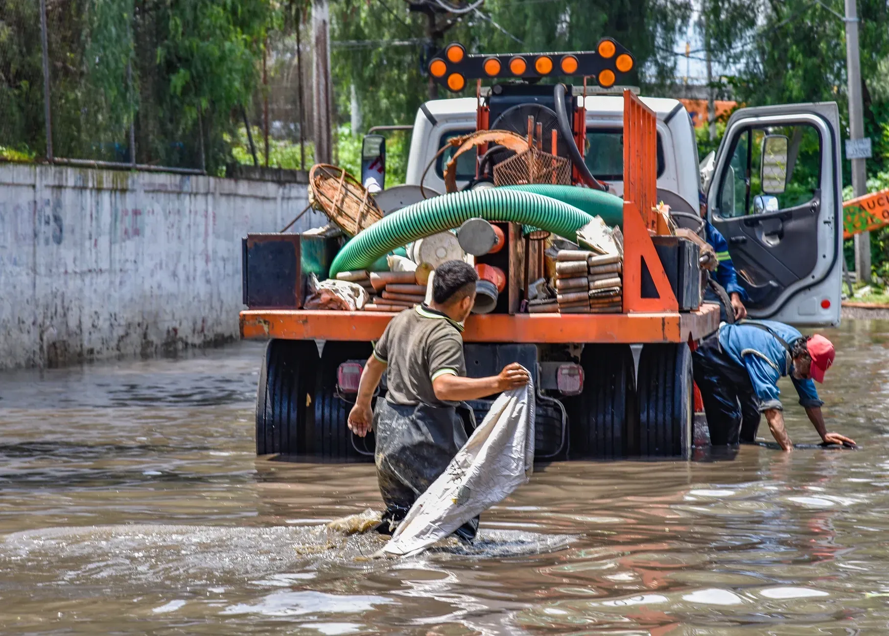 Workers loading a hose onto a truck in floodwater near a concrete wall
