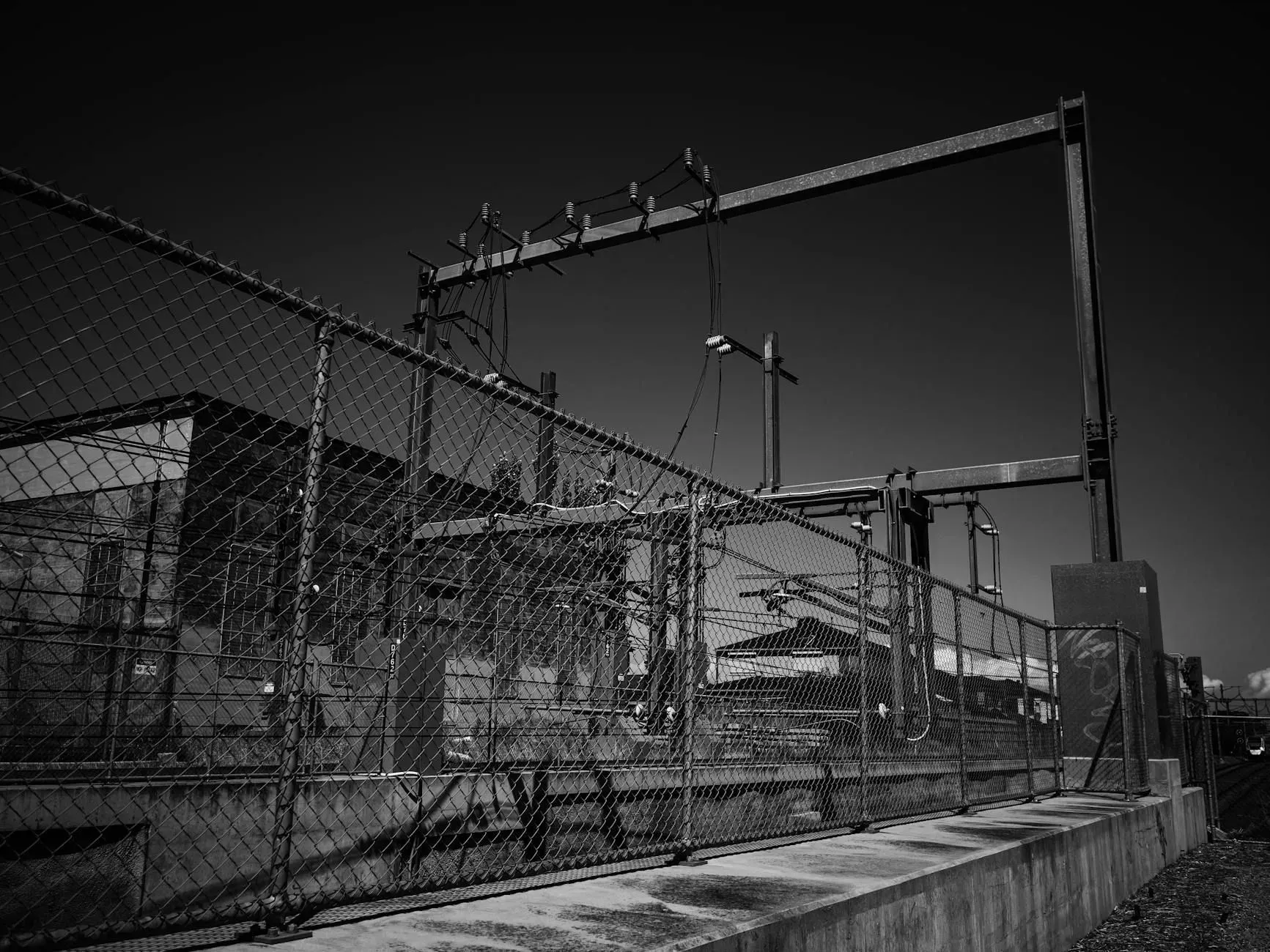 Black-and-white photo of a fenced industrial facility with overhead pipes and equipment under a dark sky
