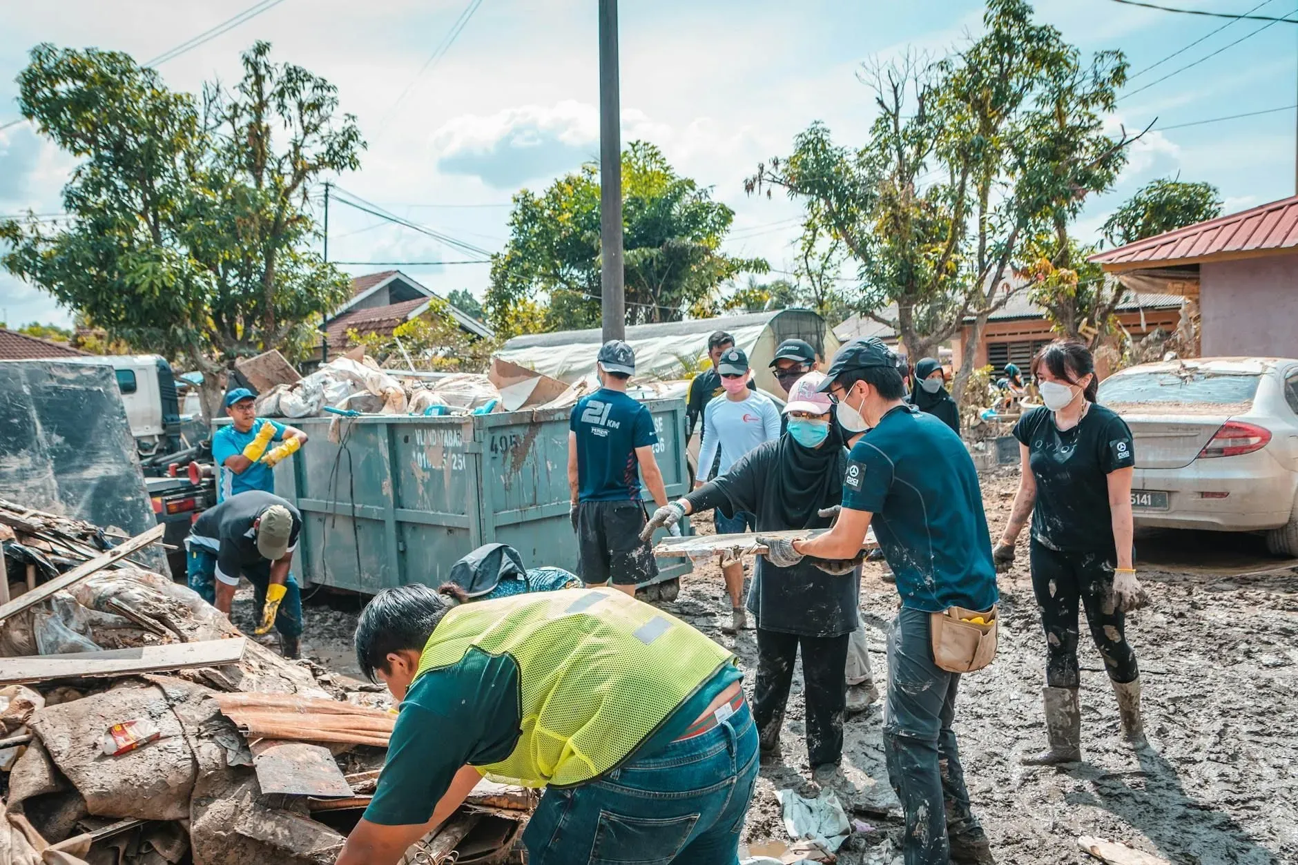 Volunteers in teal shirts clean up muddy debris beside a damaged street and parked cars.