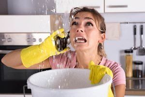 Woman in yellow gloves scrubbing with a sponge while water splashes into a bucket in a kitchen