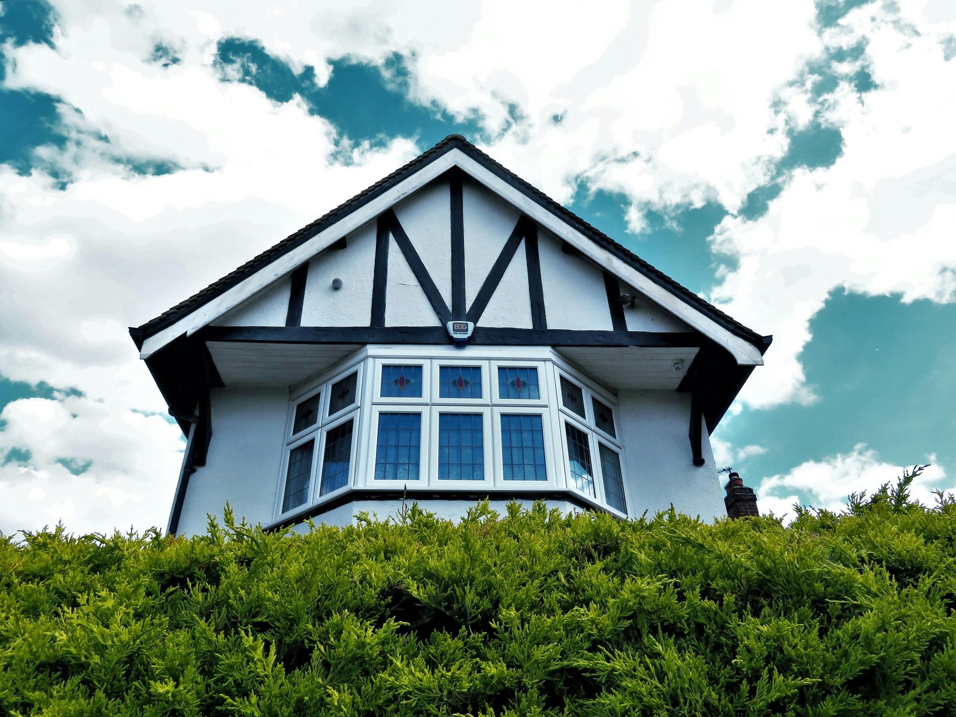 White house with black trim and a bay window, set above a green hedge, with a blue sky background.