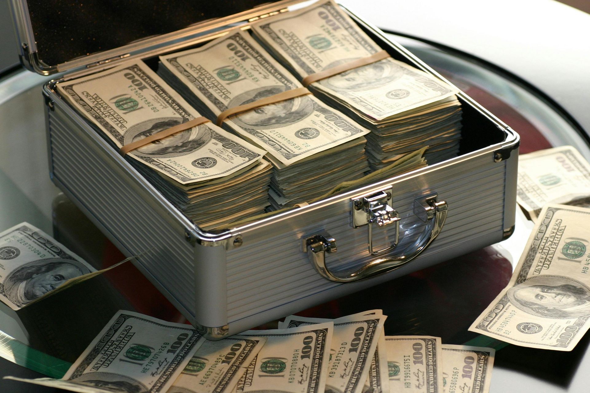 Silver briefcase overflowing with stacks of US hundred dollar bills, scattered money on surface.