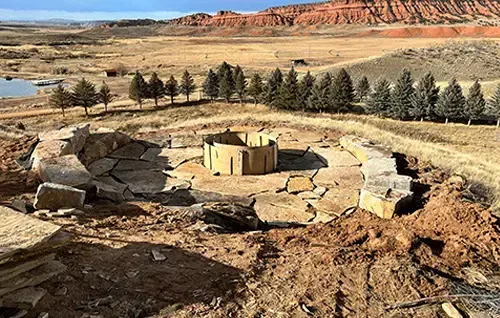 Stone circular ruin in a desert landscape with red cliffs and a line of trees in the background