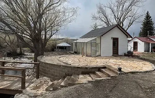 Small white shed with stone patio and steps in a bare backyard under cloudy skies