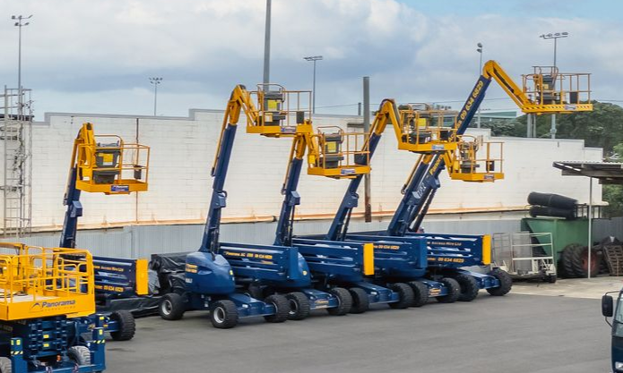 Yellow and blue boom lifts parked in a row at an industrial yard, with raised arms against a white wall.