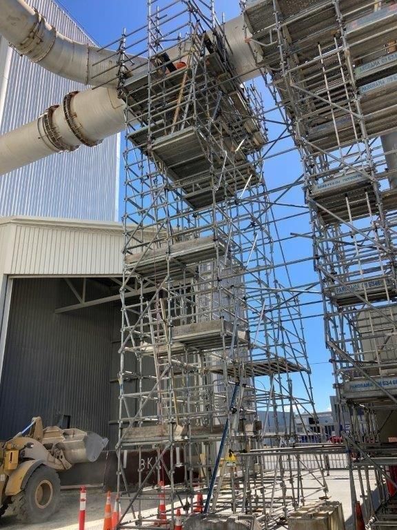 Scaffolding around industrial piping beside a warehouse, with a forklift and utility equipment below.