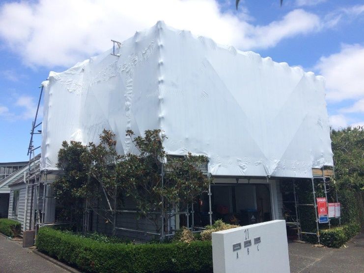 Building wrapped in white protective sheeting, with trees and hedges in front under a clear sky