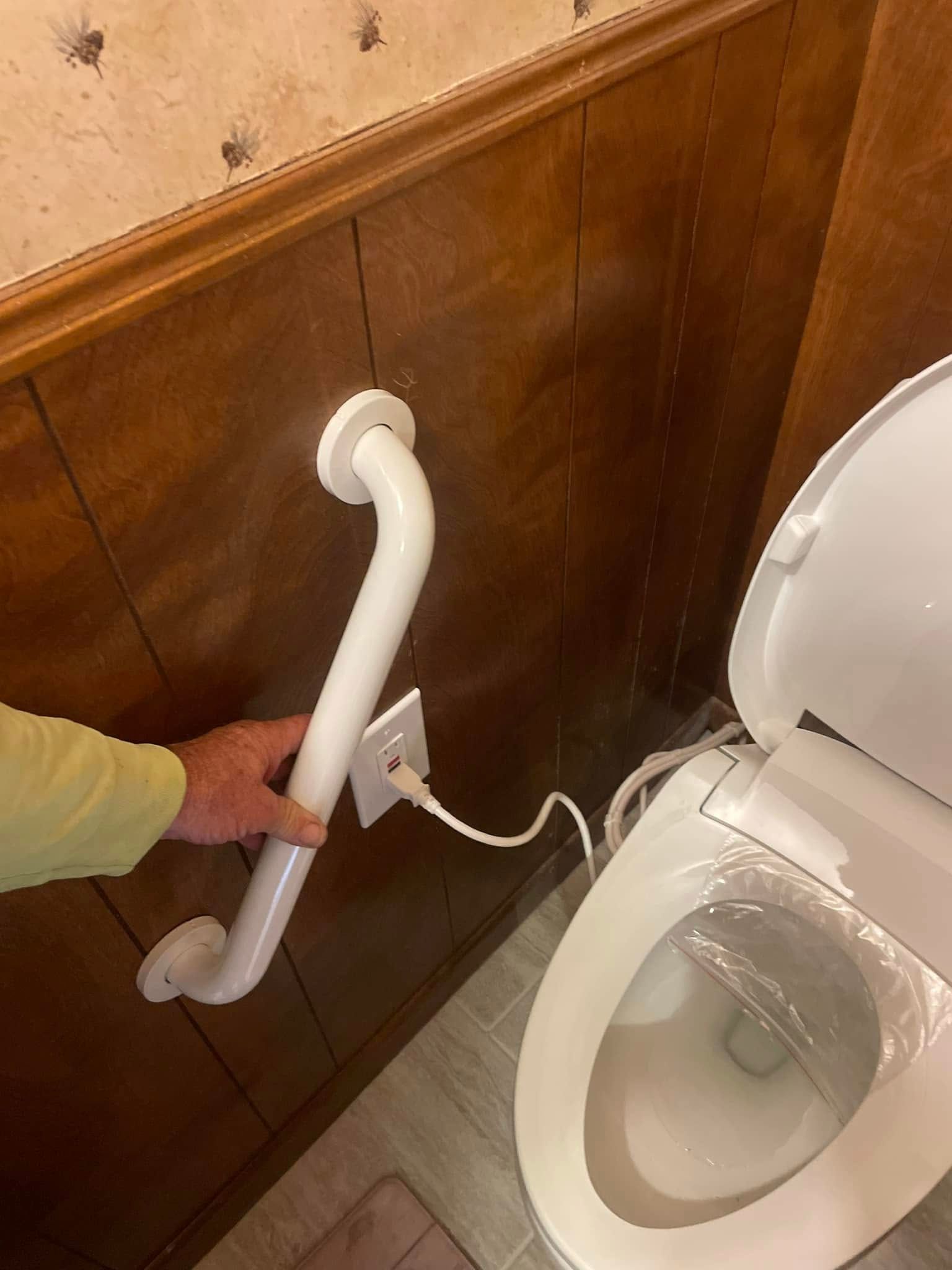 Hand using a white grab bar near a toilet and electrical outlet on wood paneling.