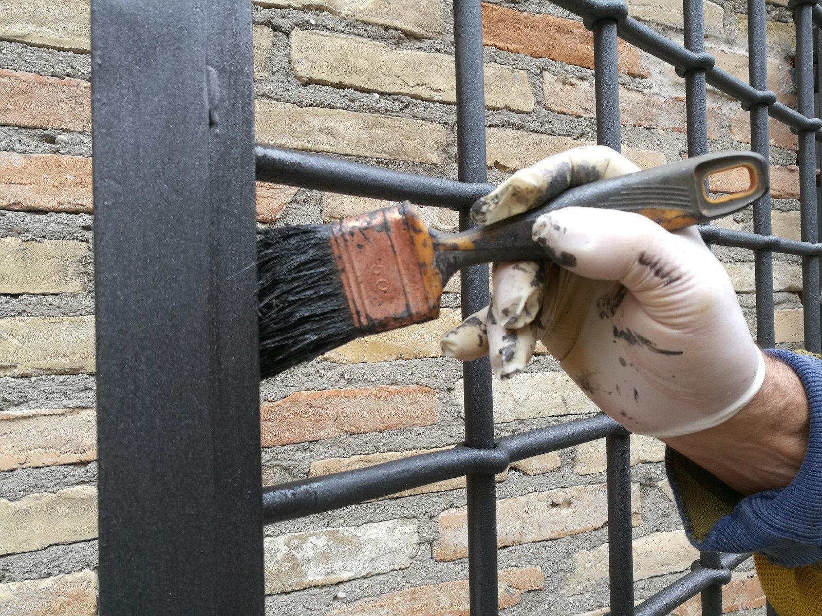 Person in gloves painting a black metal fence with a paintbrush; brick wall background.
