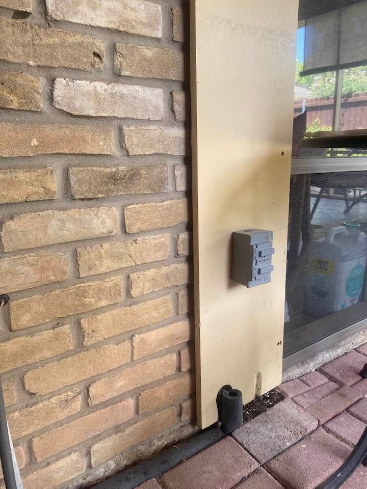 Exterior view: electrical outlet on tan panel next to brick wall and sliding glass door.