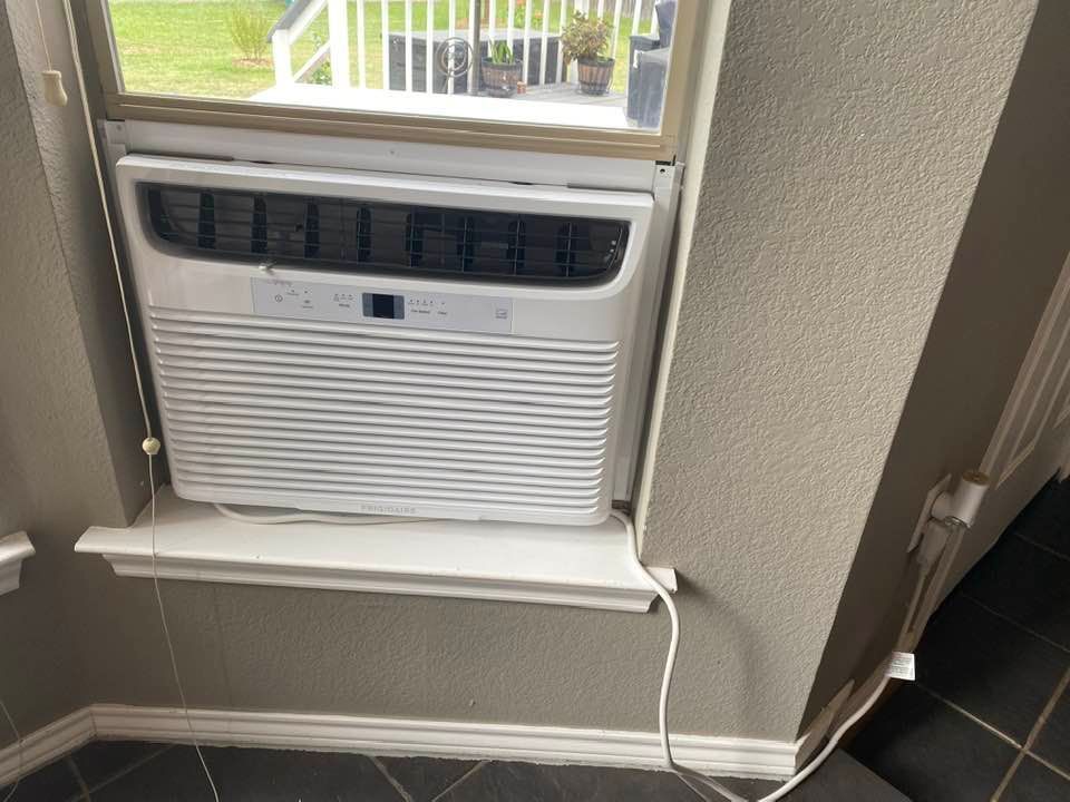 White air conditioner unit installed in a window, view of outside, set in a gray-walled interior.
