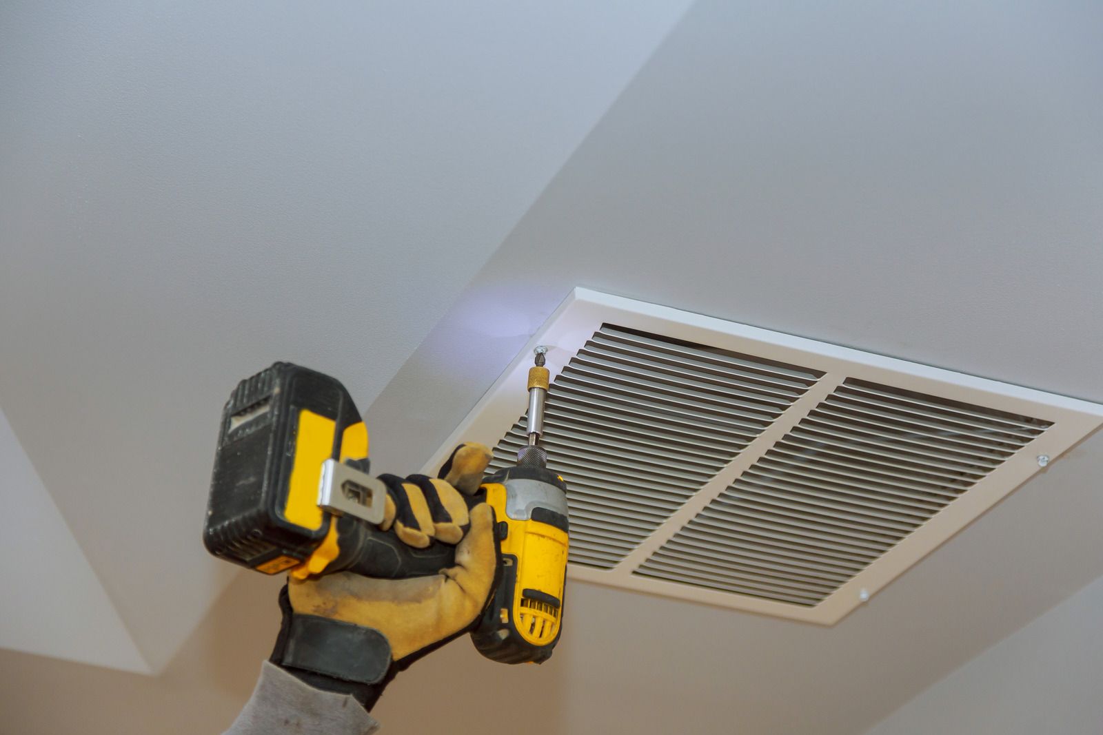 Person using a yellow and black drill to screw in an air vent cover on a white ceiling.