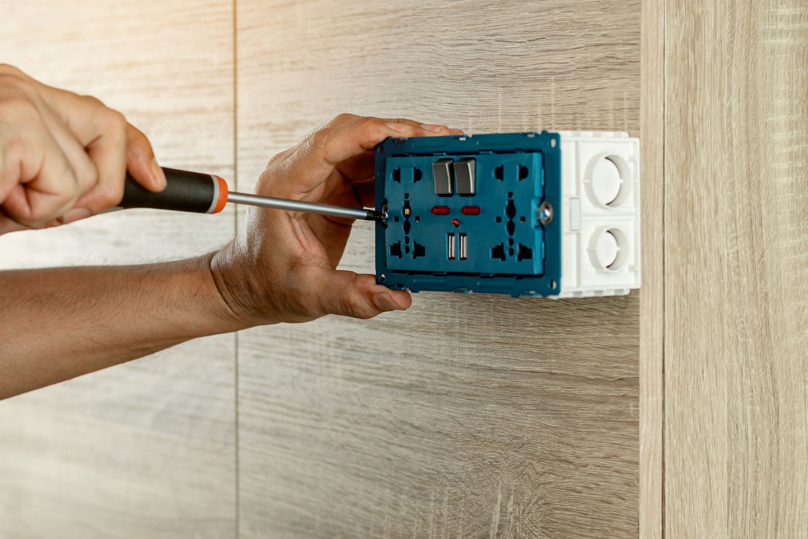 Hands using a screwdriver to install a blue electrical outlet box on a light-colored wall.