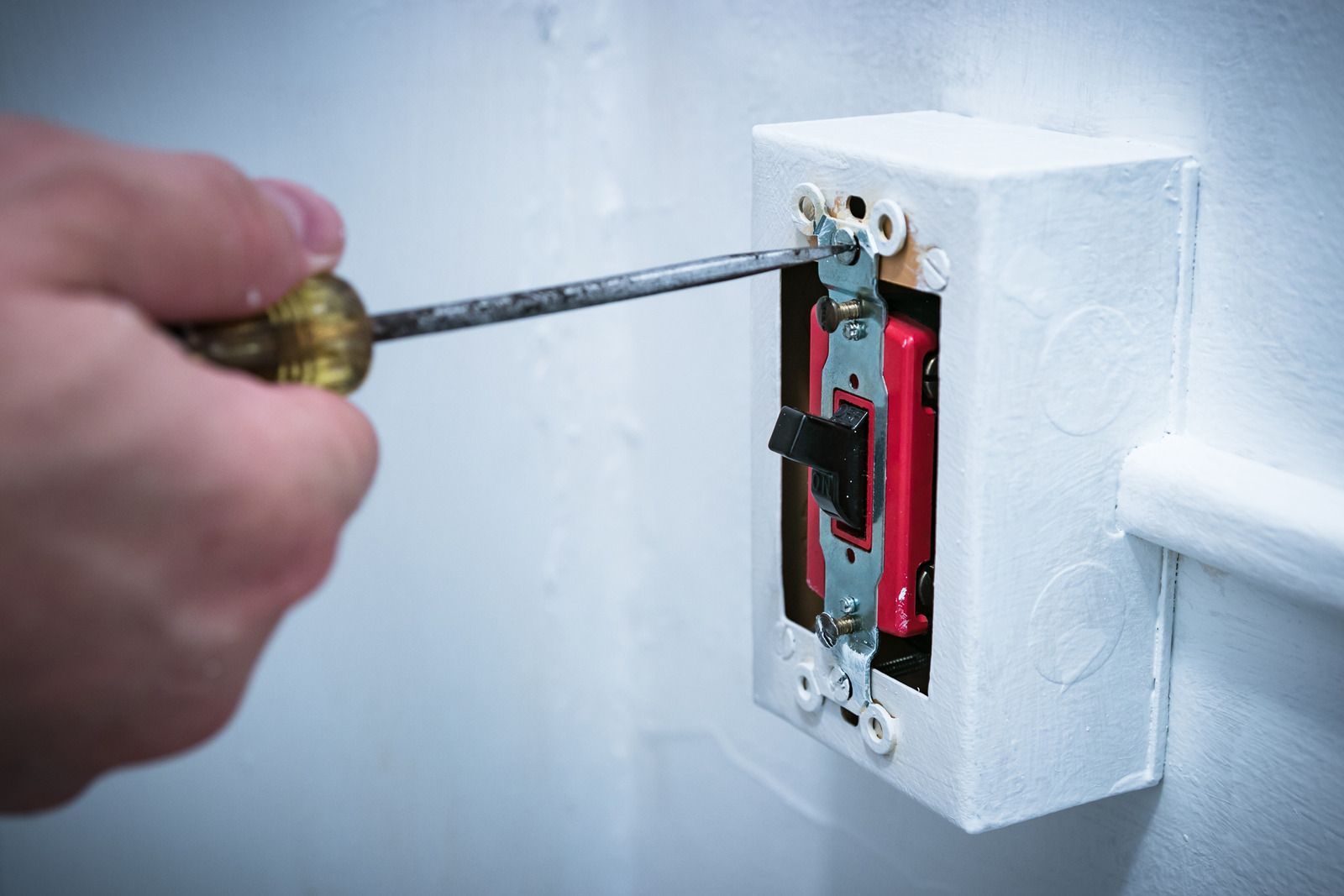 Hand using screwdriver to tighten screw on electrical switch inside white box mounted on wall.