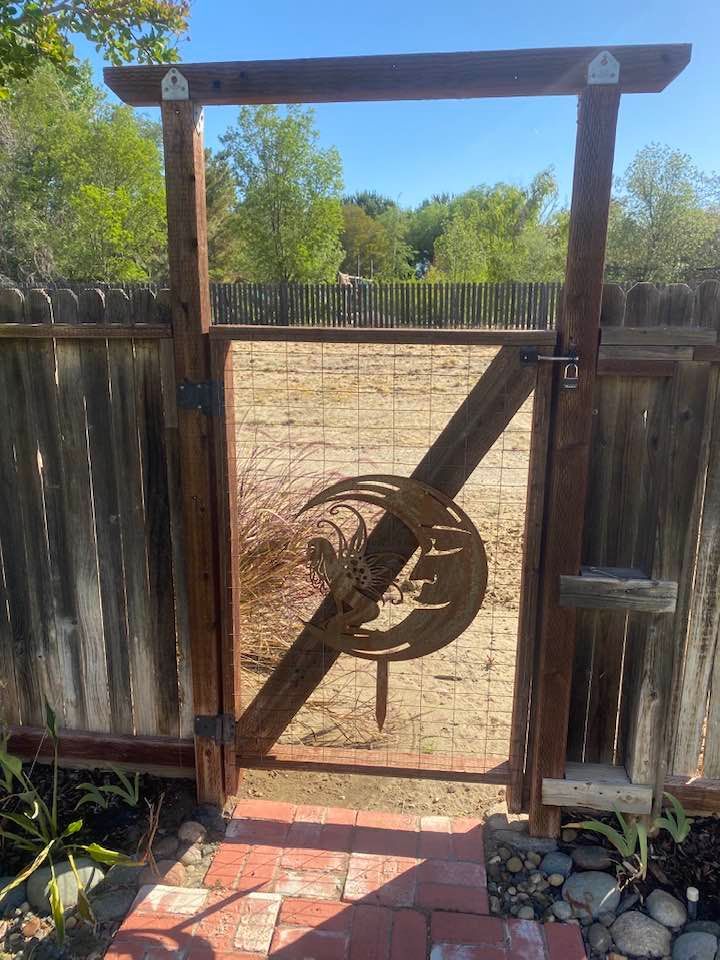 Wooden garden gate with metal art design; brick path, fenced yard, and blue sky.