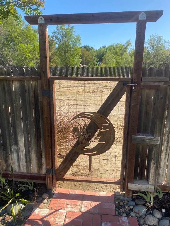 Wooden garden gate with metal art design; brick path, fenced yard, and blue sky.