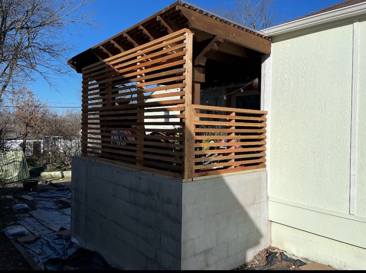 Wooden slatted gazebo on a concrete base, attached to a light yellow building.