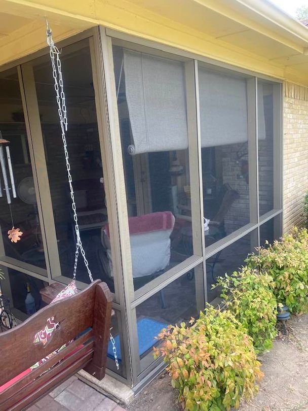 Screened porch with swing, beige trim, and rolled-up shades. Bushes along the side and brick wall.