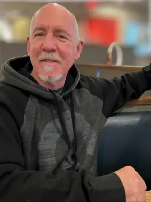 Man with a shaved head and goatee smiling, wearing a gray and black hooded sweatshirt, seated in a booth.