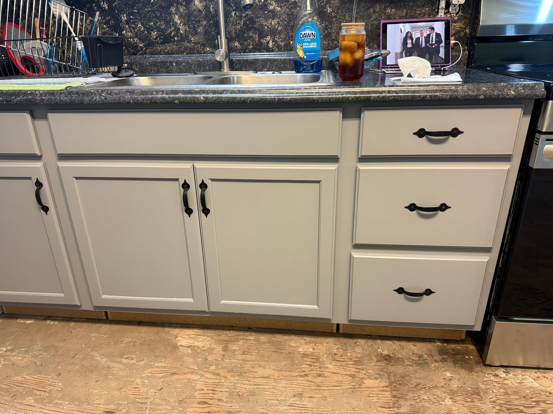 Kitchen cabinets, painted gray, with black hardware. Countertop with sink, dish soap, and a beverage.