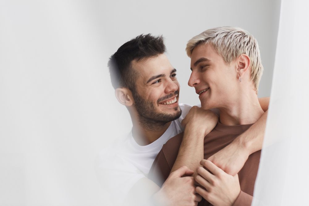 Two men embracing, smiling. One with dark hair, the other with blonde hair. Soft background, indoor setting.