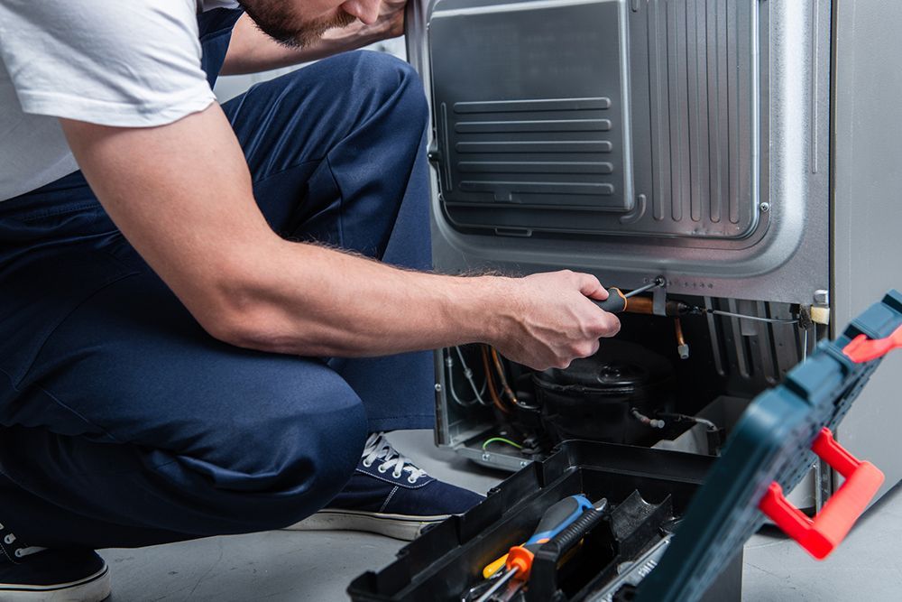 A man is kneeling down fixing a refrigerator.