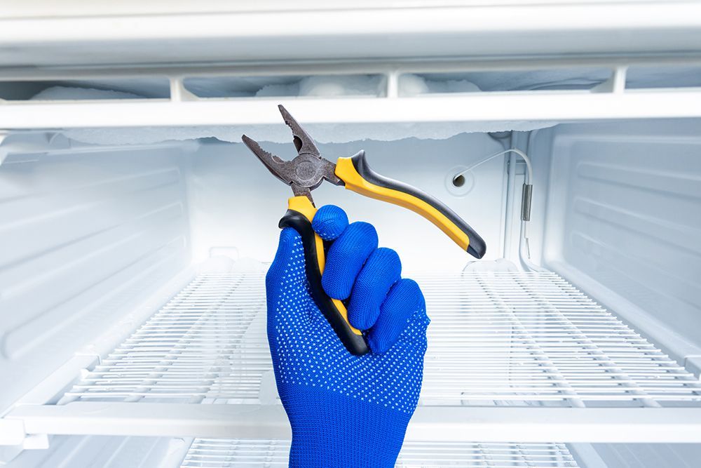 A person is holding a pair of pliers in front of an empty refrigerator.