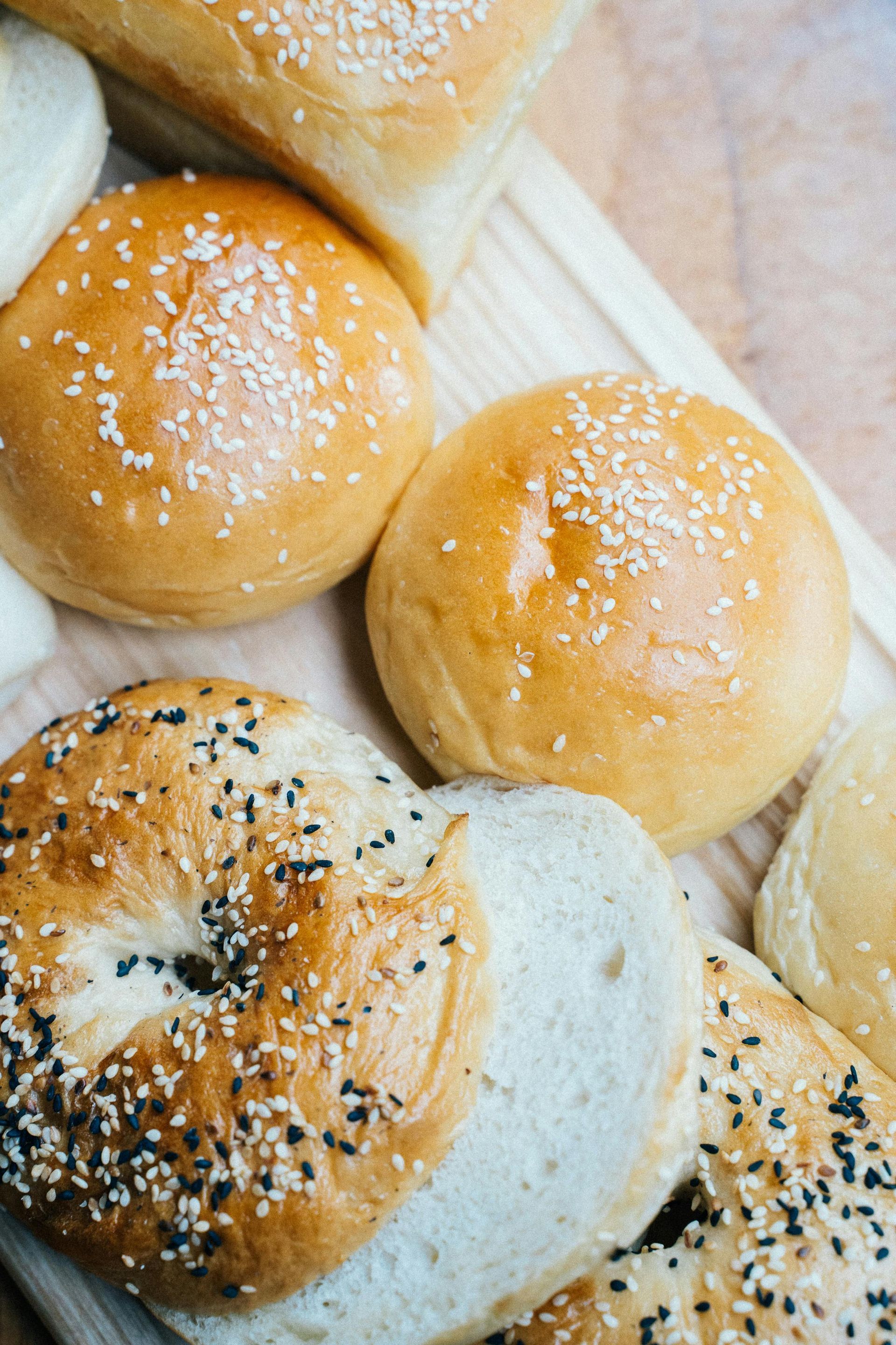 A bunch of bagels and hamburger buns on a wooden cutting board.